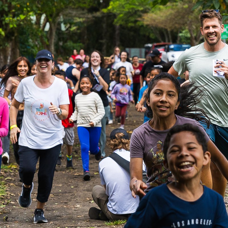 Company volunteers run alongside smiling children on a tree-lined path during a community outreach event, highlighting teamwork and giving back.