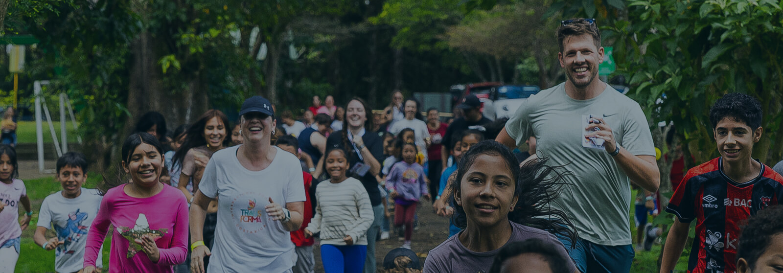Darkened Header - company volunteers run alongside smiling children on a tree-lined path during a community outreach event, highlighting teamwork and giving back.